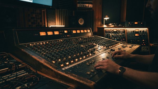 A moody, atmospheric shot of a professional audio mixing studio. Focus on a vintage-style mixing desk with warm, glowing faders and knobs. Soft, indirect lighting highlights acoustic panels on the walls. No screens or plugin UIs are visible, emphasizing t