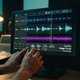 A close-up shot of the Oszillos Mega Scope plugin UI on a computer screen in a dimly lit, modern studio environment. The oscilloscope displays a complex, gated waveform, with visible rhythmic cuts and shapes. Hands are subtly adjusting a knob on the plugi