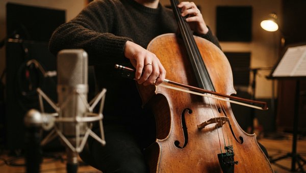 A close-up shot focusing on the hands of a musician gently bowing a cello, with a high-quality condenser microphone positioned carefully in the foreground, slightly blurred. The background shows a dimly lit, warm-toned studio space with subtle acoustic pa