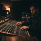 A dimly lit professional mixing studio, focusing on a large mixing desk with glowing faders and monitor speakers. No screens or plugin UIs are visible. The atmosphere is concentrated and calm, suggesting deep audio work.