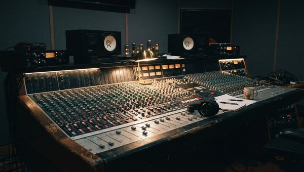 A moody wide shot of a professional recording studio, focusing on a mixing desk with faders and knobs. Soft, diffused lighting highlights the texture of the equipment. Monitor speakers are visible in the background, along with subtle acoustic paneling. No