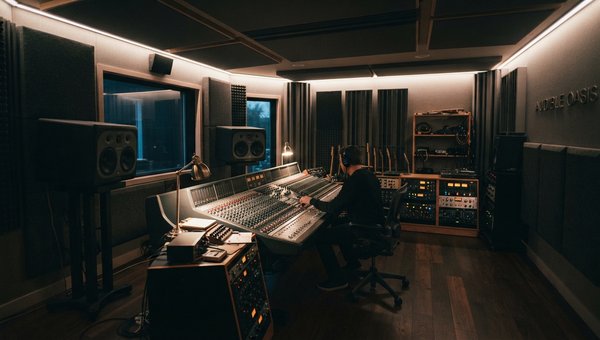 A wide shot of a professional, acoustically treated mixing studio. Soft, diffused lighting creates a focused, calm atmosphere. Acoustic panels and bass traps are subtly visible on the walls and corners. A high-end mixing desk is central, with studio monit