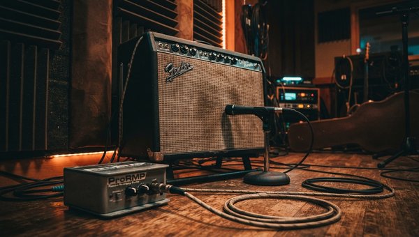 A moody, professional recording studio environment. In the foreground, a close-up of a re-amp box connected to a vintage-style guitar amplifier. A microphone is positioned in front of the amp's speaker. The lighting is soft and warm, highlighting the gear