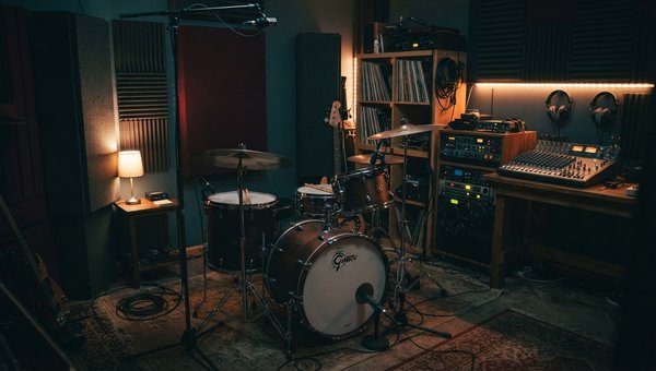 A warm, intimate shot of a compact home recording studio setup. A drum kit is positioned carefully in the center, with acoustic panels and bass traps visible in the corners and on the walls. Subtle lighting highlights the drum shells and cymbals. No compu