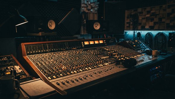 A moody, low-light shot of a professional audio mixing studio. Focus on a large mixing console with illuminated faders and knobs, monitor speakers, and acoustic panels in the background. No screens or plugin UIs are visible. The atmosphere is contemplativ