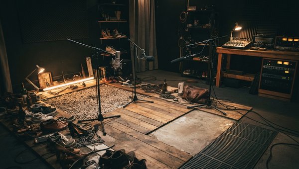 A moody, atmospheric shot inside a foley recording stage. Microphones are set up over various surfaces like wooden planks, gravel, and concrete. A foley artist's props are scattered around, but no person is visible. Soft, focused lighting highlights the t