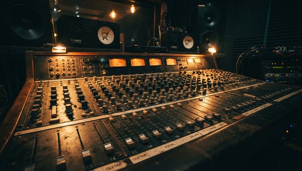A deeply moody, cinematic close-up shot of a vintage-style analog mixing desk within a professional recording studio. The intricate array of faders, knobs, and illuminated VU meters on the desk are bathed in a rich, warm golden-amber light, casting soft s